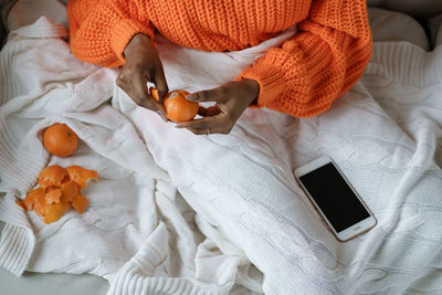 Afro woman hands peeling ripe sweet tangerine, wear orange sweater, lying in bed under the plaid