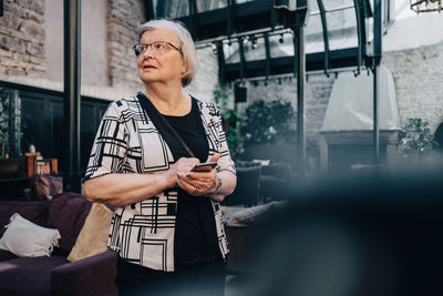 Senior female tourist looking away while holding smart phone at restaurant