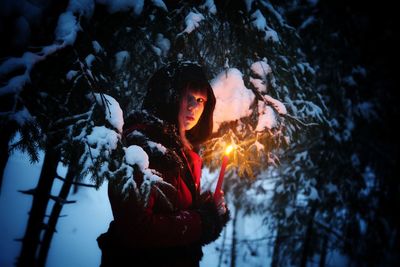 Portrait of young woman holding red illuminated candle by trees in winter