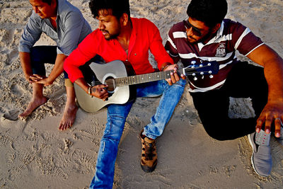 High angle view of man playing guitar while sitting with friends at sandy beach