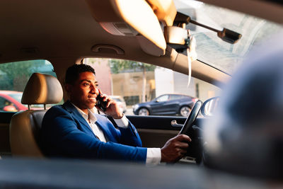 Side view of successful young african american male entrepreneur in elegant formal suit talking on mobile phone while driving car on city street