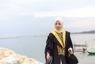 Portrait of young woman in graduation gown standing at beach against cloudy sky