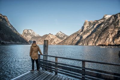 Rear view of man standing on railing by lake against sky