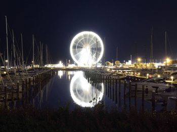 Illuminated ferris wheel by river against sky at night