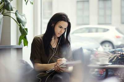 Creative businesswoman wearing headphones while writing in spiral notebook at office