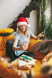 A smiling girl in a christmas hat sits on the sofa and communicates via video link with friends.