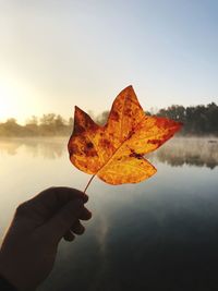 Cropped hand holding maple leaf by lake against sky during sunset