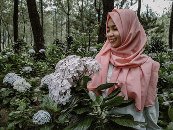 Smiling woman wearing hijab standing by flowering plants in forest