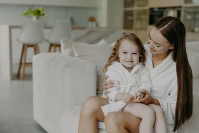 Smiling mother with daughter sitting on sofa in bathrobe