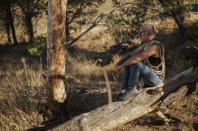 Adult man sitting by tree trunk in forest