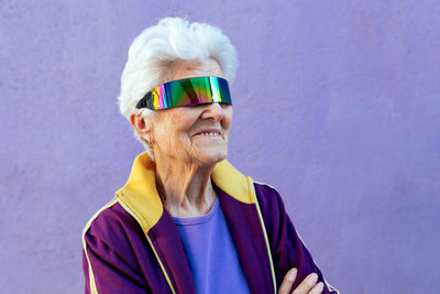 Cheerful elderly female athlete with folded arms and gray hair in sportswear and blindfold on violet background