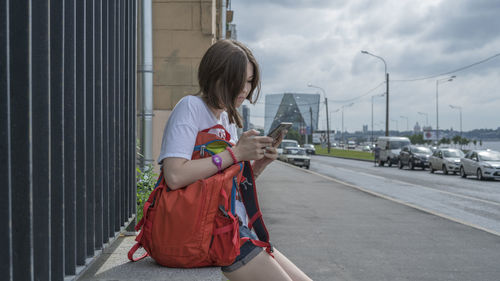 Young woman using smart phone while sitting on roadside in city