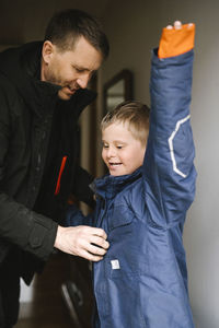 Father helping son with down syndrome to wear jacket standing at home