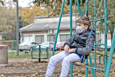 Little boy swinging at the playground while wearing face mask due to coronavirus pandemic.