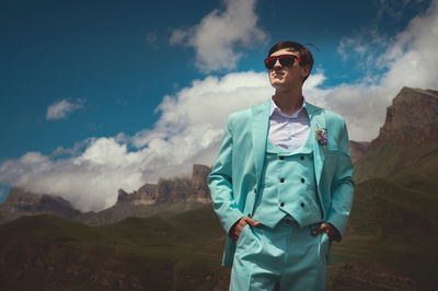 Groom in a suit stands against a background of mountains and clouds in summer. a man in a turquoise