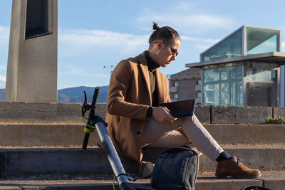 Businessman sitting on the stairs of the city using tablet with the electric scooter leaning behind 