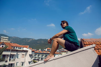 Man in the green t-shirt shorts and sunglasses sitting on the roof of a clay orange tile