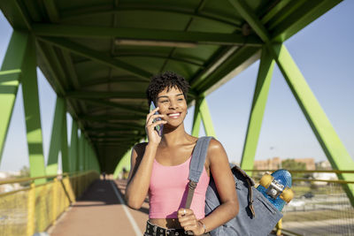 Smiling woman with backpack talking on smart phone at bridge