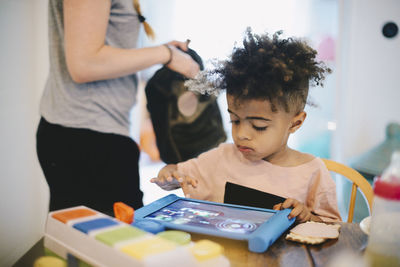 Boy using digital tablet at table while mother standing in background
