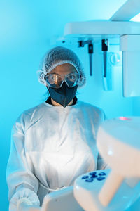 Professional female doctor in medical uniform and protective mask with goggles looking at camera while using modern diagnostic equipment in hospital