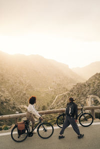 Multiracial male and female friends looking at mountains while wheeling bicycles on road