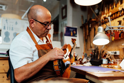 Side view of mature male luthier in apron and glasses sitting on chair and holding restored violin while working in workshop