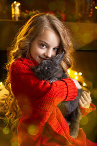 A little blonde girl with a gray rabbit in her arms next to a christmas tree decorated with garlands
