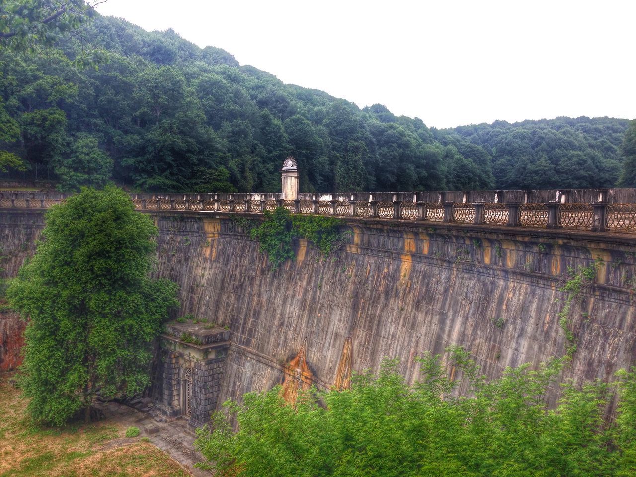 mountain, built structure, architecture, tree, water, clear sky, building exterior, tranquility, tranquil scene, nature, scenics, beauty in nature, river, bridge - man made structure, mountain range, connection, growth, plant, outdoors, sky