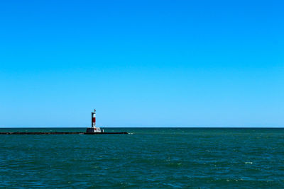 Lighthouse by sea against clear blue sky