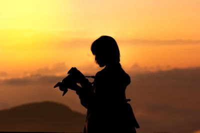 Silhouette woman holding camera against sky during sunset