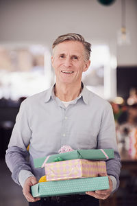 Portrait of senior man holding stacked gift boxes while standing at home