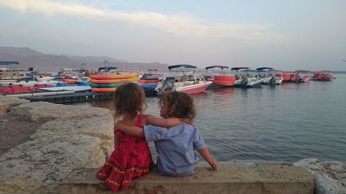 Rear view of siblings with arms around on retaining wall against boats