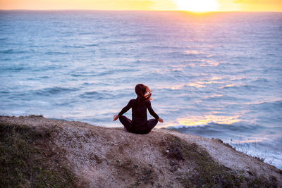 Man sitting on rock by sea against sky during sunset