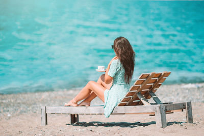 Woman holding coffee cup sitting on chair at beach