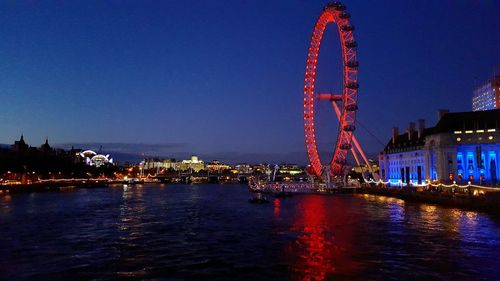 Illuminated ferris wheel by river against sky at night