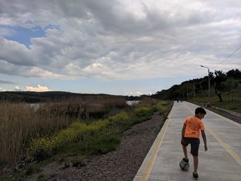 Rear view of boy playing soccer on footpath against cloudy sky