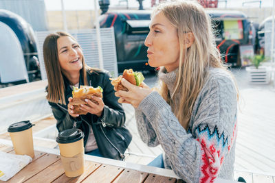 Two attractive women eat street fast food and have fun chatting while sitting at a table outside.