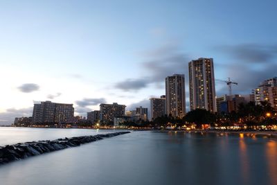 Illuminated buildings by river against sky in city