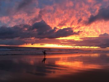 Silhouette man with surfboard standing at beach against sky during sunset