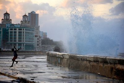 Boy running away from sea waves splashing on retaining wall in city during sunset