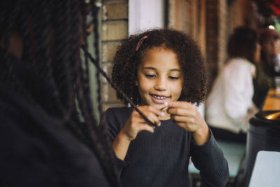 Smiling daughter with curly hair braided hair of mother while sitting at restaurant