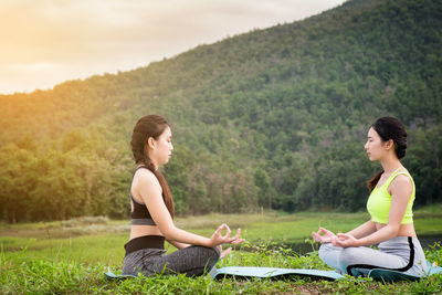 Side view of female friends doing yoga on field against mountain