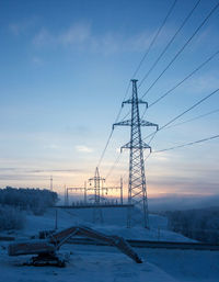 Electricity pylon on snow covered field against sky during sunset