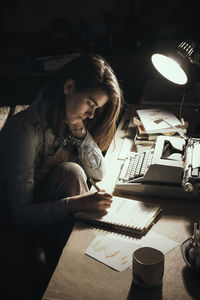 Woman writing by lamp at desk with typewriter