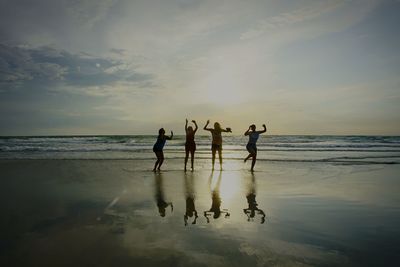 Friends jumping on shore at beach against sky during sunset