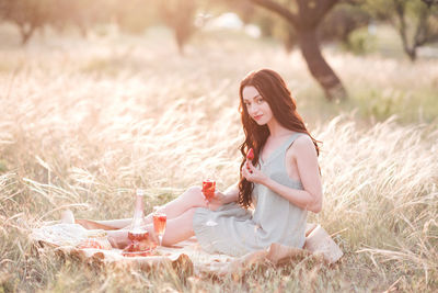 Happy woman having romantic picnic in meadow with wine bottle and fruits outdoors. 20s.