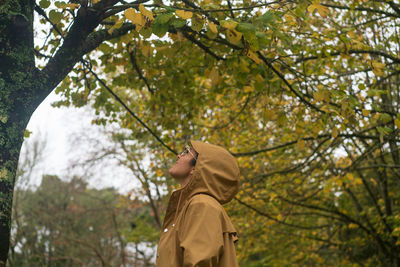 Caucasian woman with yellow rain jacket looking at autumn fall yellow leaves tree on a rainy day