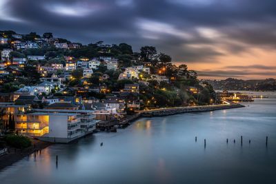 Illuminated buildings by sea against sky at sunset