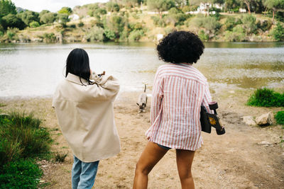 Back view of unrecognizable female friends with dog and skateboard standing near river and admiring nature during camping trip together in summer countryside