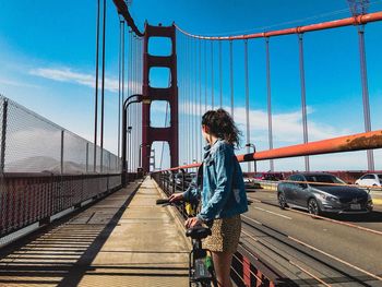 Side view of woman standing on footbridge against blue sky